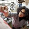 A student holds a paintbrush, looking up at a canvas as she begins to paint, her focus directed toward her work. The brush is poised, ready to add to the artwork in front of her.