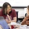 A professor and two students are seated at a table in class, engaged in conversation. Their computers are open in front of them, with the group discussing ideas or working collaboratively on a project.