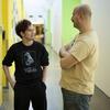 Two students are talking and smiling in a hallway, standing still and engaged in conversation. Their relaxed body language and friendly expressions suggest an easygoing, enjoyable exchange.