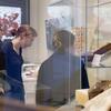 Two students are examining books in a glass display case, likely in a museum space. They appear focused and engaged as they explore the displayed items, with the museum's surroundings adding a thoughtful and educational atmosphere.