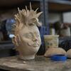 A clay sculpture of a head with spiky hair and a mustache rests on a table in a ceramics studio. The sculpture showcases intricate details, with the texture of the clay adding depth to the facial features and hair.