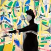 A woman dressed in all black with her face covered in silver sequins extends her arms. Behind her is a vibrant multicolor wall featuring shades of blue, yellow, pink, and green.