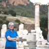 Jane DeRose Evans stands outside with her arms crossed, looking at the camera. Behind her is an outdoor landscape featuring Roman architectural ruins.