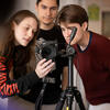 Three students stand behind a camera on a tripod. The girl on the far left holds the lens, carefully adjusting the shot, while the others stand nearby, observing and assisting with the setup.