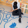 A woman sits on the ground, carefully placing pieces of blue tape on the floor to create a design.