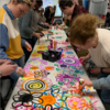 A group of students gathers around a long table, collaboratively painting colorful flowers onto its surface.