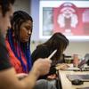 A student and professor sit at a table, looking at the student's laptop and discussing what's on the screen. They are engaged in conversation, with the professor offering feedback or insights while the student listens attentively.