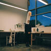 Ollie Goss stands in a studio space wearing a goose hat, with a large window behind them. The scene is set at dusk or dawn, with soft, natural light filtering through the window, creating a calm and creative atmosphere.