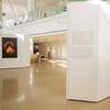 A photo exhibition area with photos neatly hung on white walls. Natural light pours in from windows above, illuminating the space and highlighting the displayed images.
