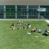 A group of students sits in a circle outside in Tyler's courtyard, with a few others hanging out to the right of them. The sun is shining, the grass is green, and the atmosphere feels warm and relaxed.
