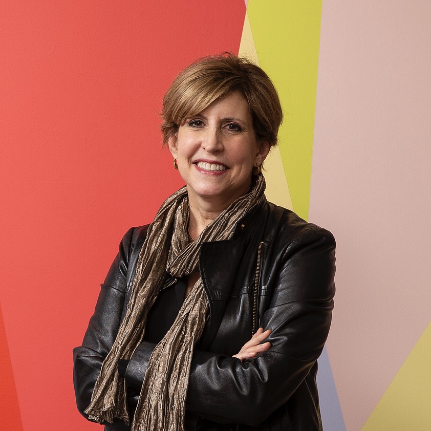 Susan Cahan stands in front of a multicolored wall with her arms crossed, smiling at the camera.