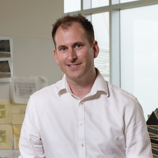 Christopher McAdams standing in an architecture studio, looking at and smiling for the camera.