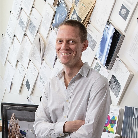 Andrew John Wit smiling, leaning on a desk with his arms crossed in a Tyler architecture studio.