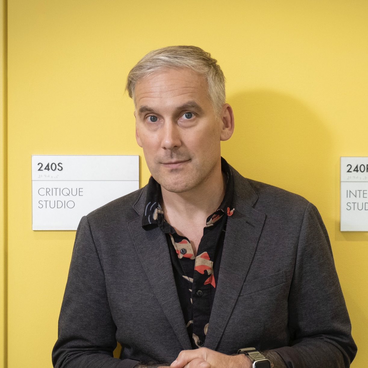 Matt Curtius stands in front of a yellow wall, with room number signs for two design classrooms at Tyler on each side of him.