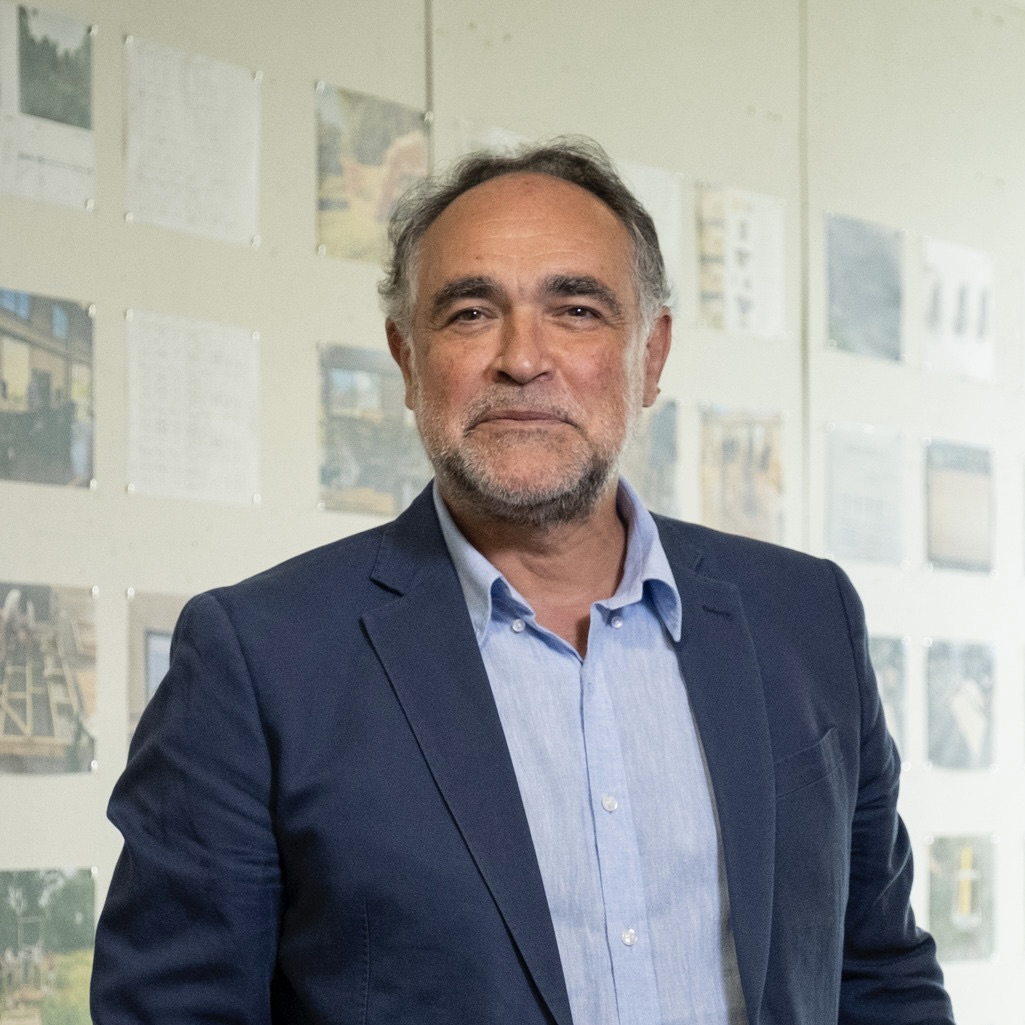 Mauricio Bertat standing and looking at the camera, with photographs of architecture projects displayed behind him.