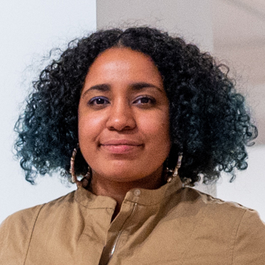 Woman with curly black and dark green hair, wearing a light brown button-up shirt and hoop earrings, looking directly at the camera with a neutral expression.