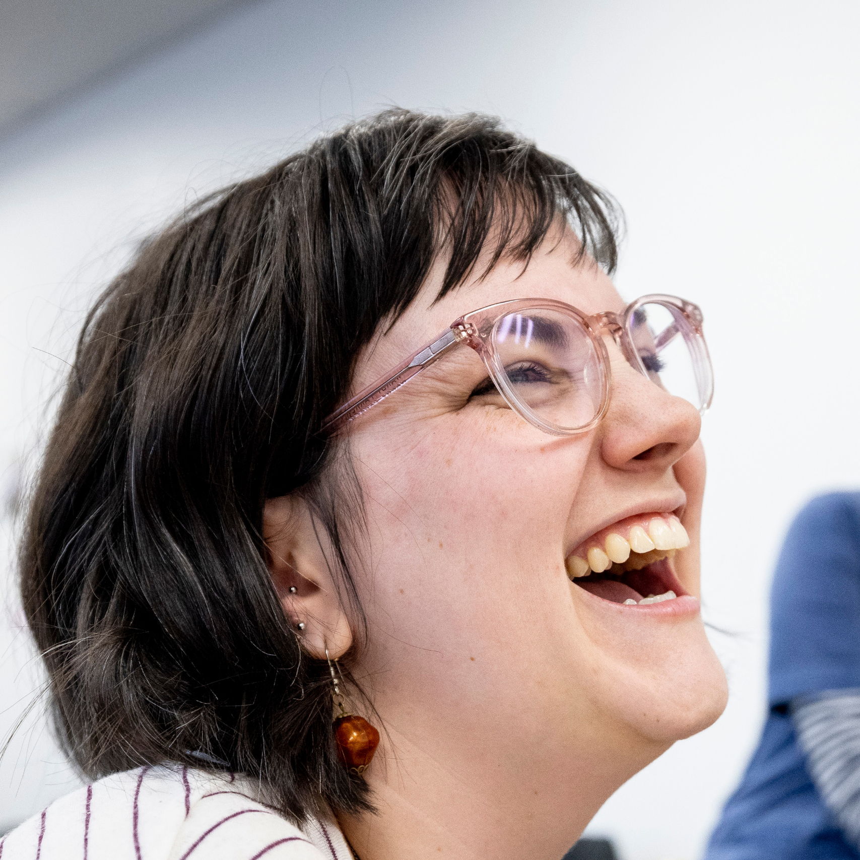 lose-up of a person laughing, wearing large light pink glasses and amber bead earrings.