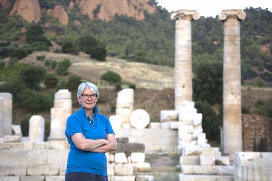 Jane DeRose Evans stands outside with her arms crossed, looking at the camera. Behind her is an outdoor landscape featuring Roman architectural ruins.