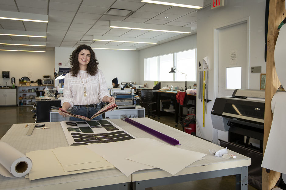 Mia Culberston stands in a Tyler design studio, holding a book in her hands and smiling at the camera.