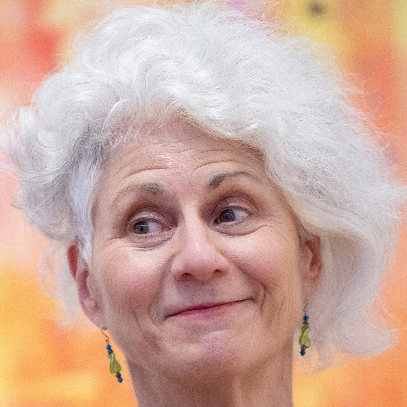 Close-up portrait of an elderly woman with wavy white hair and dangly green and blue earrings.